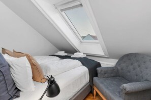 Une chambre avec un puits de lumière, un lit avec des oreillers, une table de chevet avec une lampe et un fauteuil gris.