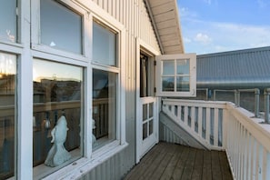 Una terraza de madera con barandilla blanca, una estatua y un edificio con ventanas grandes y techo de metal.