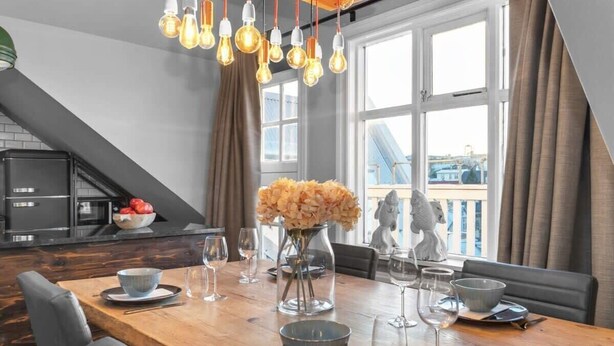 Een eetgedeelte met een houten tafel, stoelen, een vaas met bloemen en hangende lampjes.