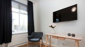 A room with a window, a chair, a desk, a flat-screen TV, and a vase with flowers.