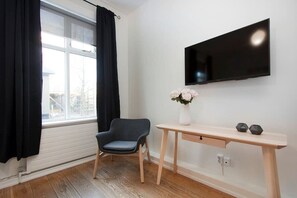 Una habitación con ventana, una silla, un escritorio, un televisor de pantalla plana y un jarrón con flores.