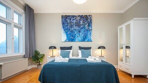 Un dormitorio con una cama, dos mesitas de noche, una lámpara auxiliar, una ventana, una planta y un armario.