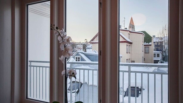 Una ventana con barandilla blanca, una orquídea en maceta y vistas de edificios nevados y el campanario de una iglesia.