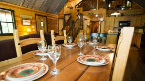 A wooden dining table set with plates and wine glasses, surrounded by wooden chairs in a rustic interior with a staircase and kitchen visible in the background. - Crazy Horse is a charming Hochatown Cabin with all the Amenities. (Broken Bow)