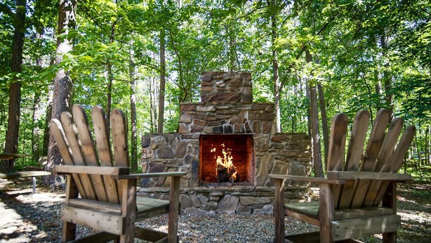 A stone fireplace with two Adirondack chairs in a forest setting. - Crazy Horse is a charming Hochatown Cabin with all the Amenities. (Broken Bow)
