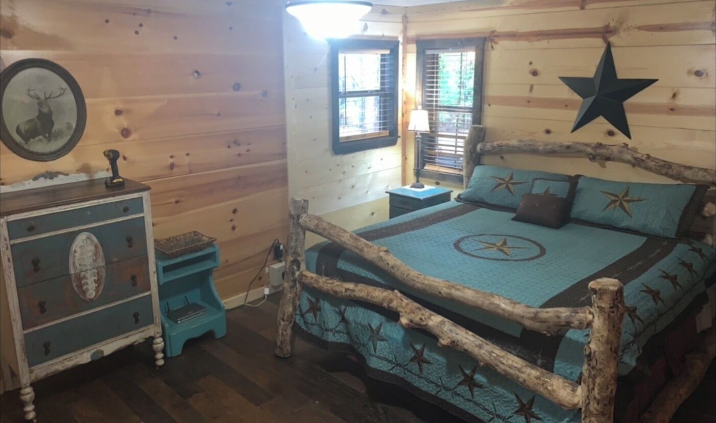 A rustic cabin-style bedroom with a bed framed by a natural log, a dresser with a decorative mirror, and a window with blinds.