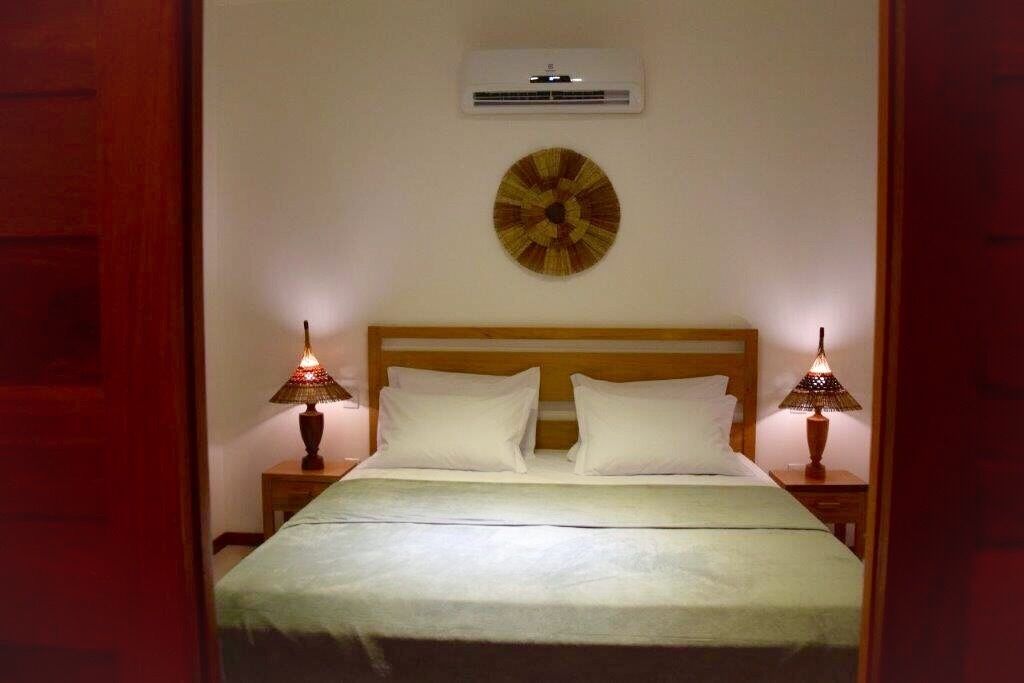 A neatly made bed with white linens, two bedside lamps, a wall-mounted air conditioner, and a decorative wall piece.