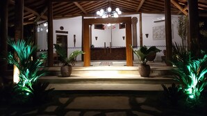 A covered outdoor area with a wooden ceiling and a central light fixture. There are two large potted plants on either side of a central pathway leading to a door. - Carapitangui Pousada (Maraú)