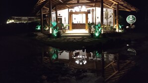 A thatched-roof structure with a well-lit entrance, surrounded by potted plants and a reflective water surface. - Carapitangui Pousada (Maraú)