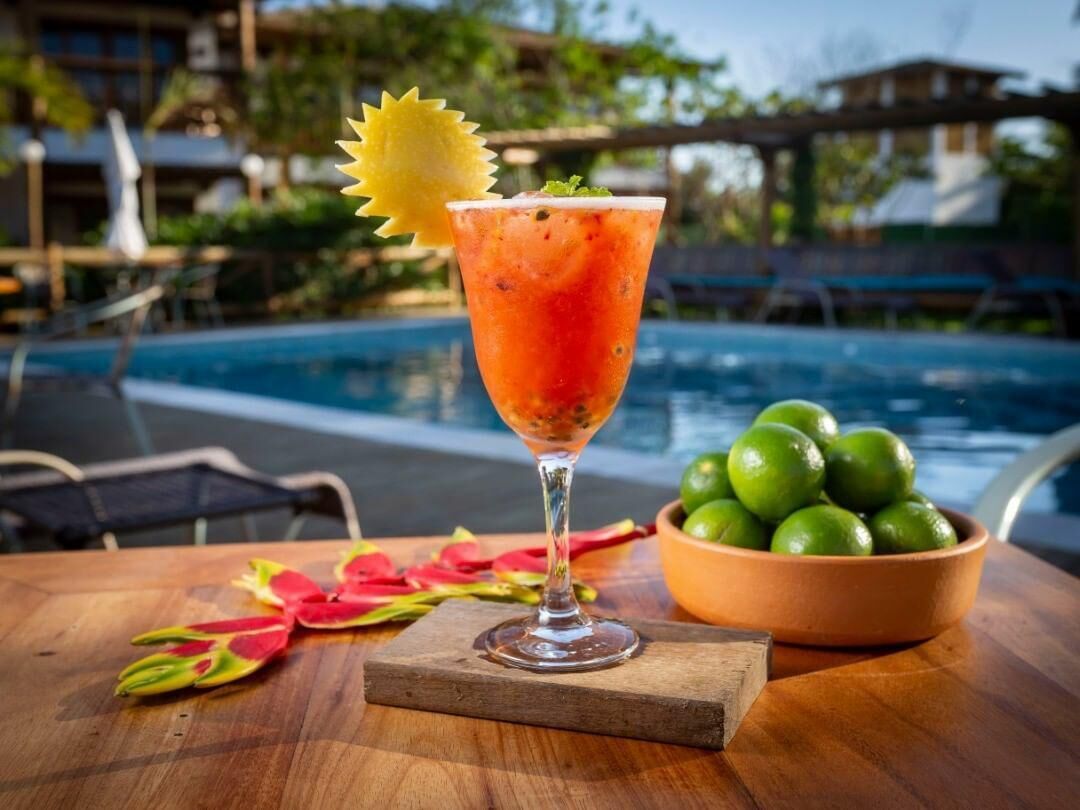 A cocktail with a garnish, a wooden table, a bowl of limes, and a pool in the background.