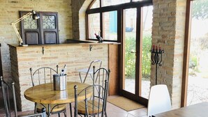 A rustic dining area with a brick wall, wooden table, metal chairs, and a large window with a view of greenery. - Villa Bella Rosa (Fratte Rosa)