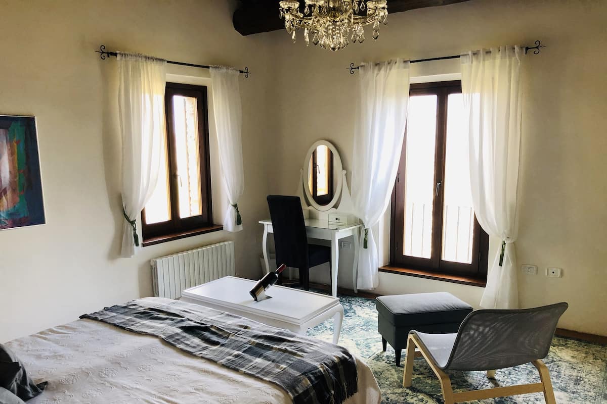 Une chambre à coucher comprenant un lit, un bureau, une chaise, un miroir et deux fenêtres avec des rideaux.