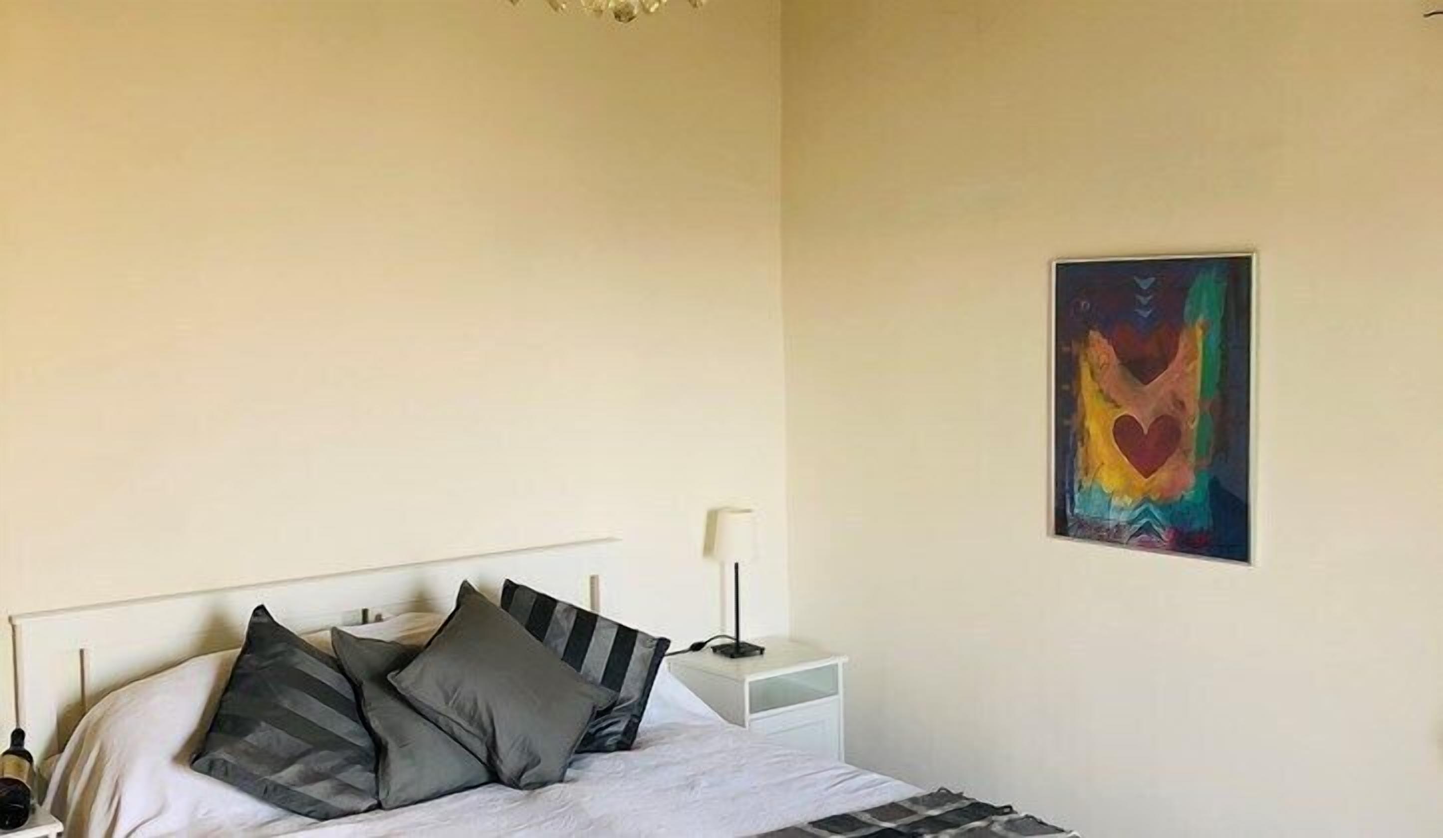 Un dormitorio con cama, almohadas, una mesita de noche, lámpara y un cuadro abstracto y colorido en la pared.