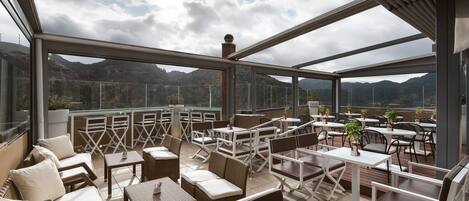 Una terraza exterior con mesas y sillas, barandilla de vidrio y vistas a montañas.