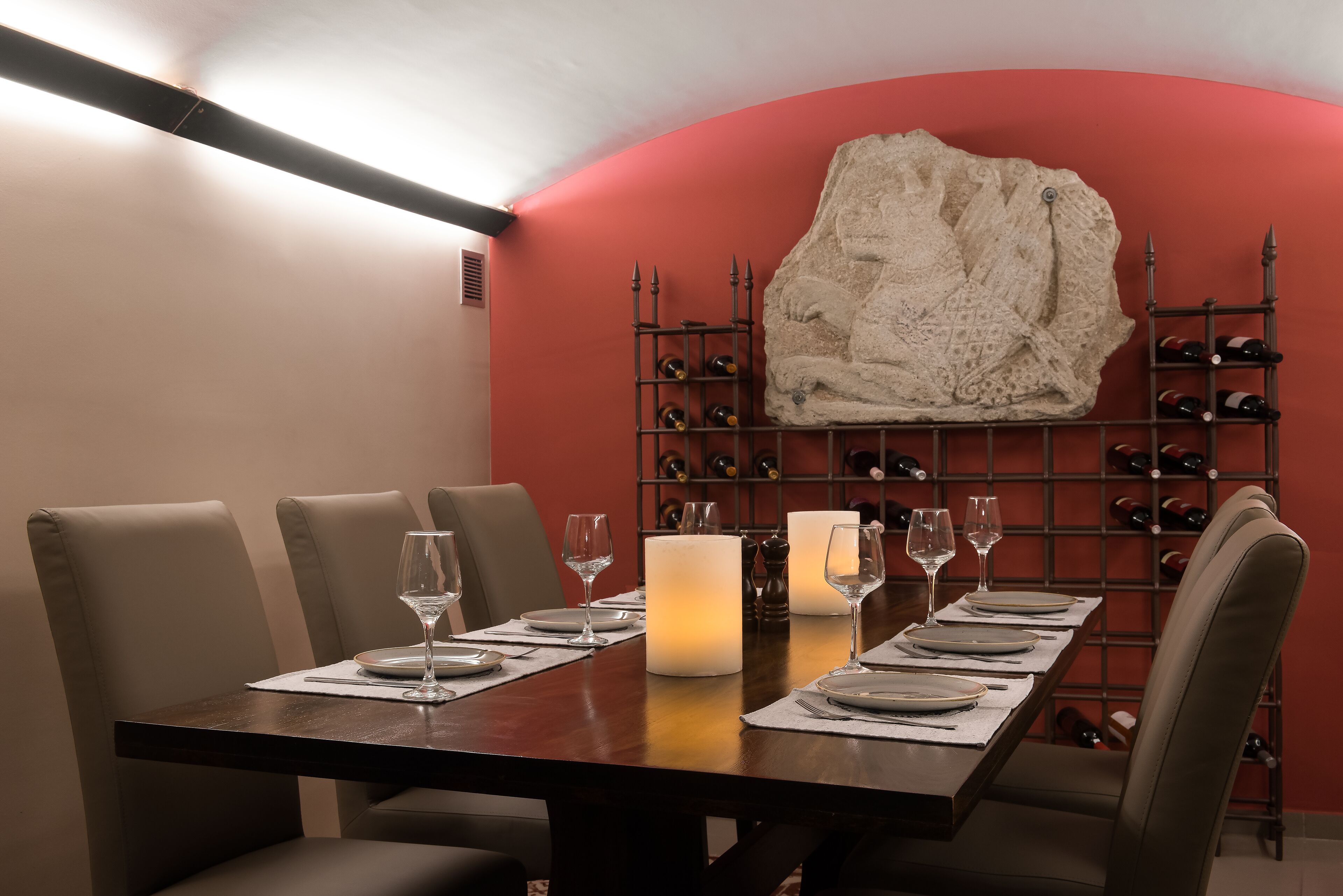 A dining area with a long table set for four, surrounded by chairs, with a wine rack and bottles in the background.