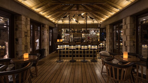 A wooden bar area with stone walls and a high ceiling. There are multiple bar stools and tables, and the bar is stocked with various bottles and glasses. - Hotel Kentrikon (Gortynia)