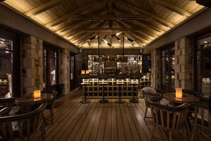 Un espace bar en bois, avec des murs en pierre et un plafond haut. On y trouve de nombreux tabourets et tables, et le bar est bien garni de bouteilles et de verres de toutes sortes.