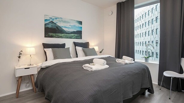 A modern hotel room with a large bed, a nightstand with a lamp, a view of a cityscape through a window, and a painting on the wall. - Sif Apartments Reykjavik - 2 Bedrooms with balcony (Reykjavík)