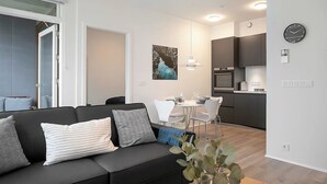 A modern living room with a black sofa, a dining area with a white table and chairs, and a kitchen with dark cabinets and a clock on the wall. - Sif Apartments Reykjavik - 2 Bedrooms with balcony (Reykjavík)