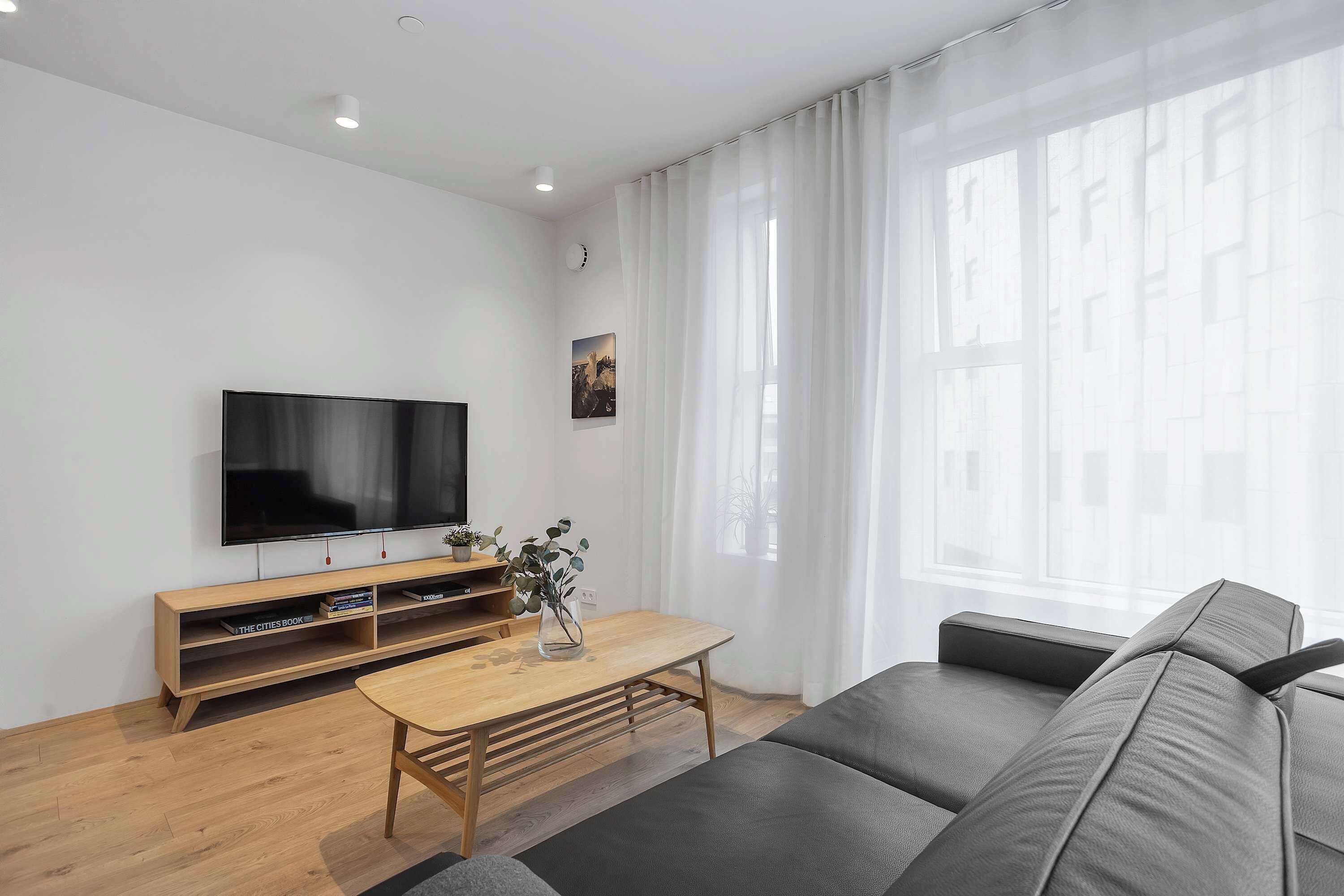 A modern living room with a flat-screen TV, wooden furniture, and a large window with sheer curtains.