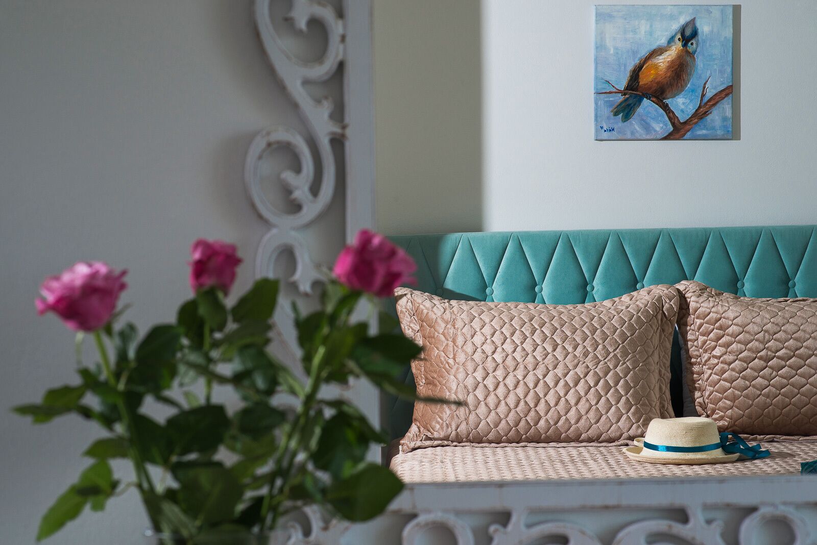 A room with a teal upholstered headboard, a quilted beige pillow, a straw hat, and a painting of a bird on a branch.
