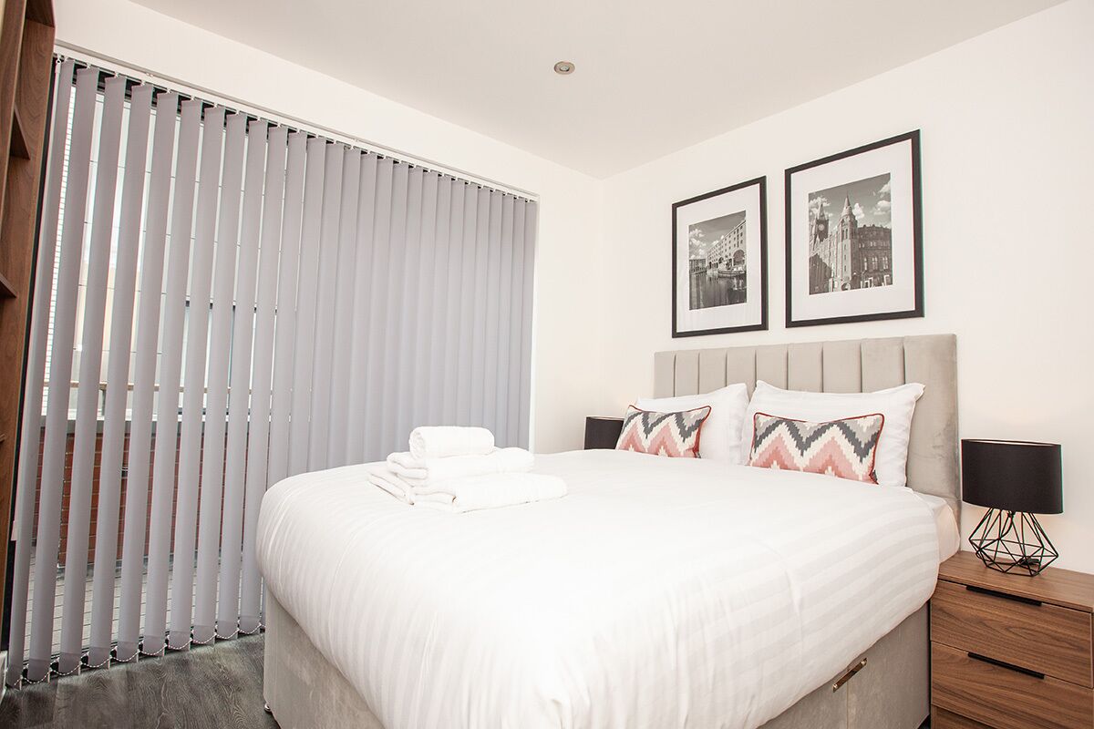 A neatly made bed with white linens, a headboard with two framed pictures, and a bedside table with a lamp.