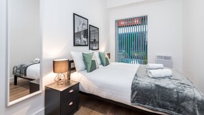 A modern bedroom with a large bed, a bedside table with a lamp, a mirror, and two framed pictures on the wall. - SGT Pepper’s Apartments (Liverpool)