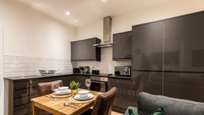 A modern kitchen with dark cabinetry, a wooden dining table, and a sofa with green cushions. - SGT Pepper’s Apartments (Liverpool)
