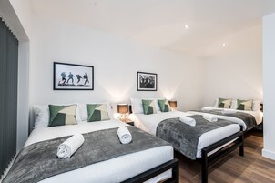 Una habitación de hotel con dos camas, cada una con una manta gris y una toalla enrollada. Hay lámparas de pared y cuadros enmarcados sobre las camas.