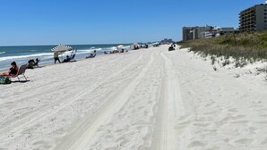 Ein Strand mit blauem Himmel, einem sandigen Pfad und Gebäuden im Hintergrund.