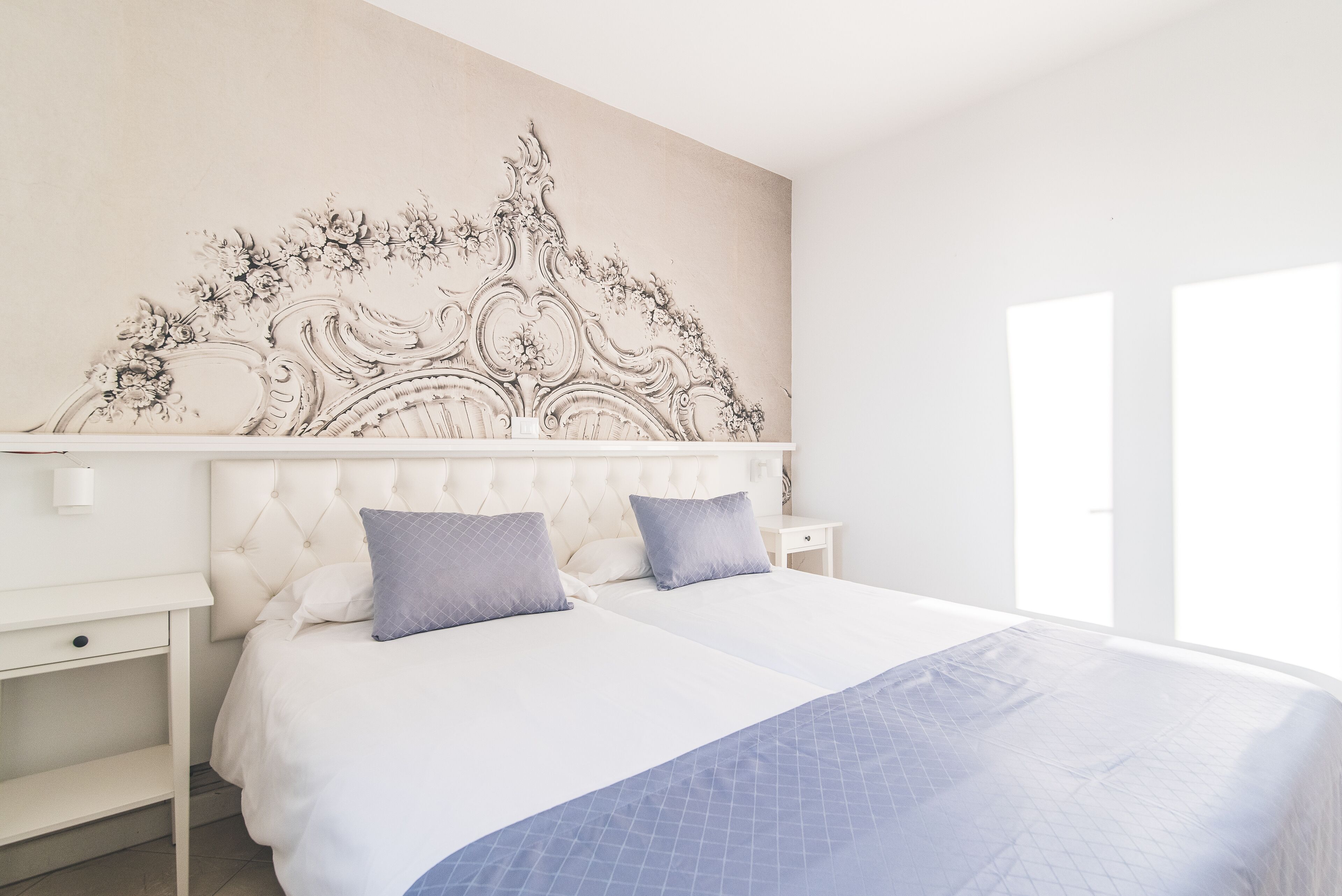 A bedroom with a tufted headboard, two beds with blue bedding, a nightstand, and a decorative wall mural.
