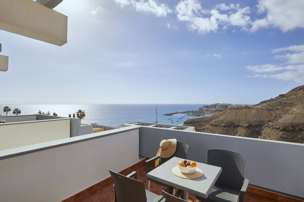 A balcony with a table, chairs, and a bowl of fruit. The view includes the sea and a distant coastline.