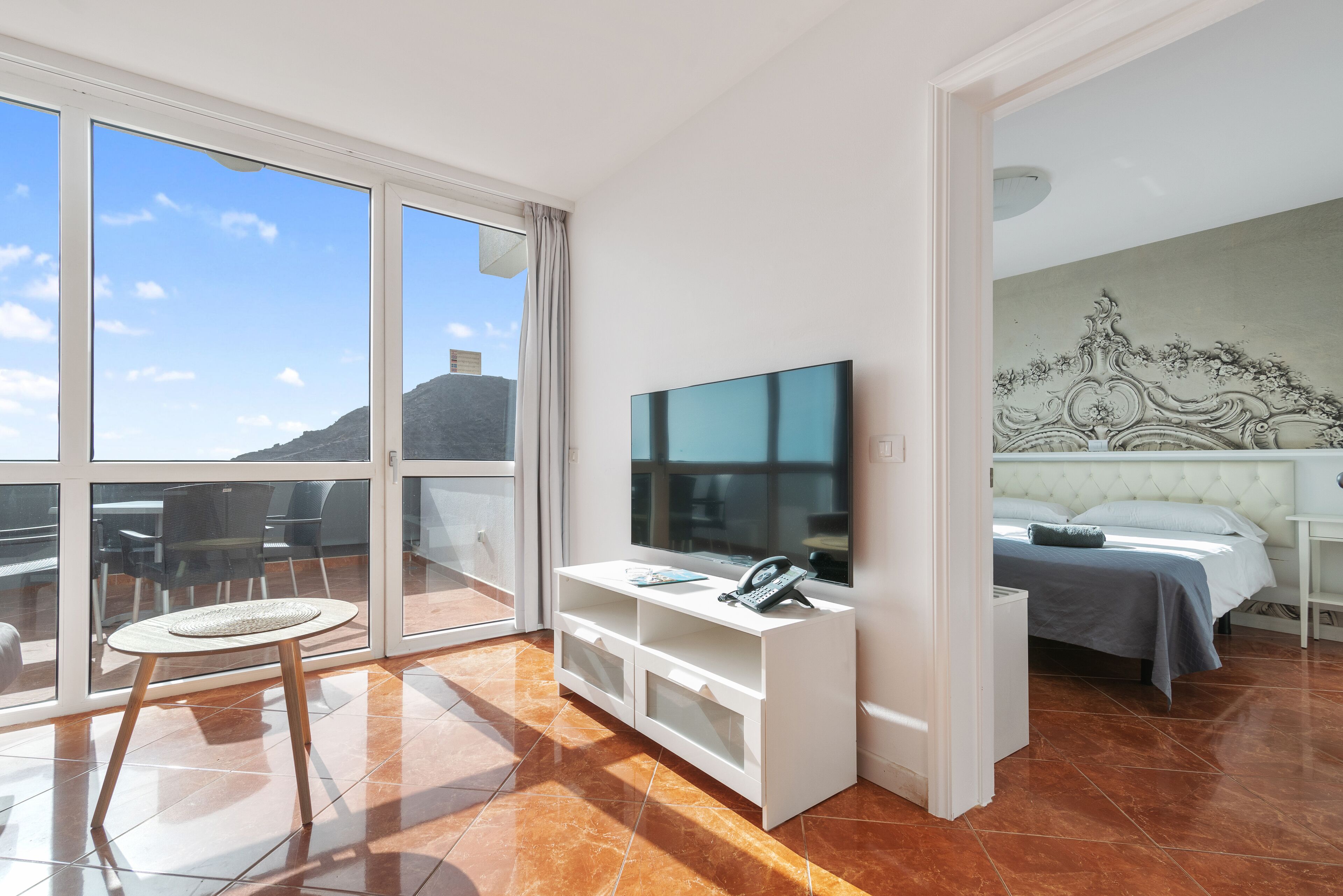 A hotel room with a large window, a flat-screen TV, a bed, and a small table with a chair.