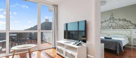 A hotel room with a large window, a flat-screen TV, a bed, and a small table with a chair.