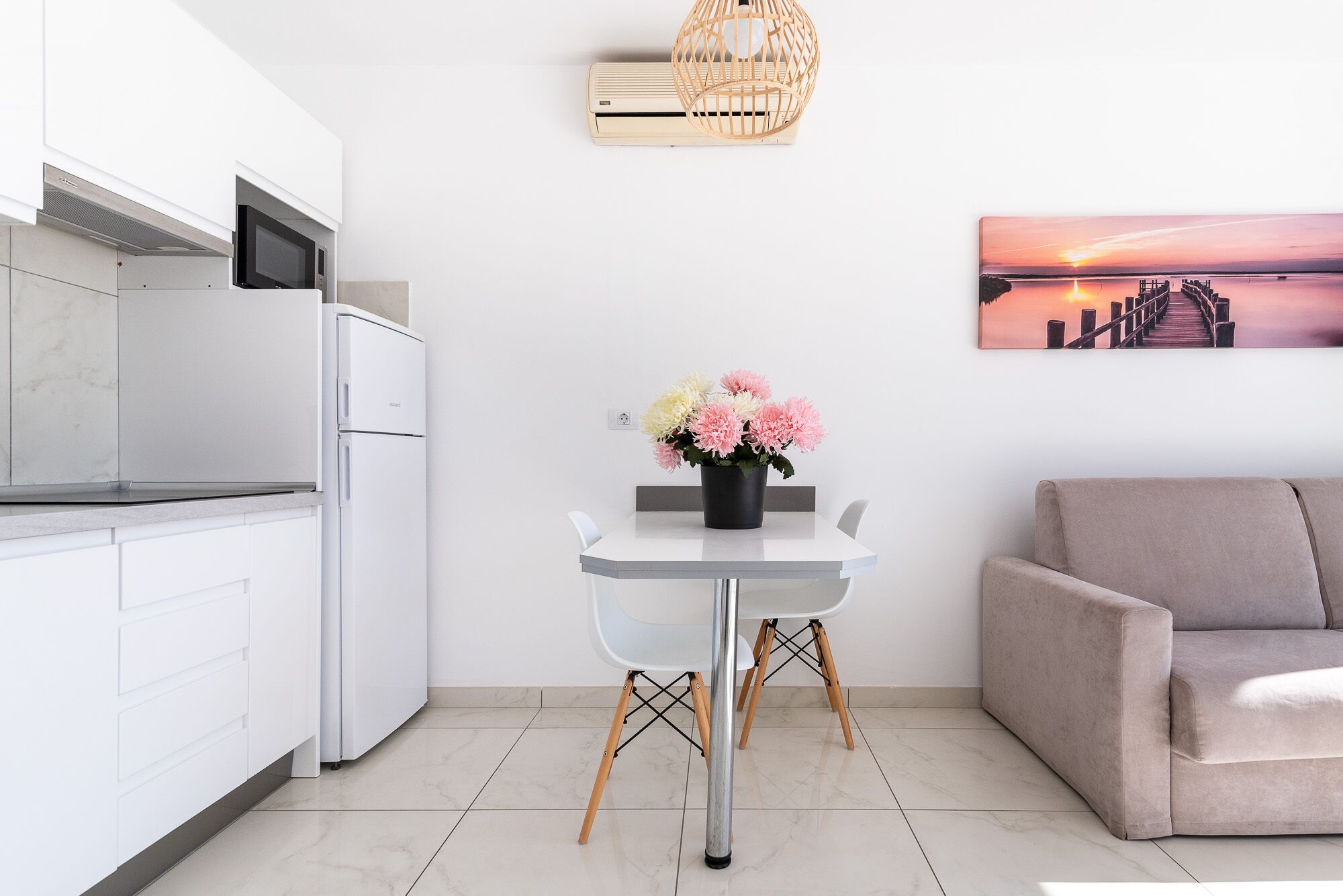 Une cuisine moderne avec des meubles blancs, un micro-ondes encastré et un réfrigérateur en acier inoxydable. On y trouve une table à manger blanche ornée d’un vase noir contenant des fleurs, deux chaises blanches et un canapé beige dans l’espace salon. Une cage à oiseaux en bois est accrochée au mur, et un tableau représentant un coucher de soleil est exposé au-dessus du canapé.