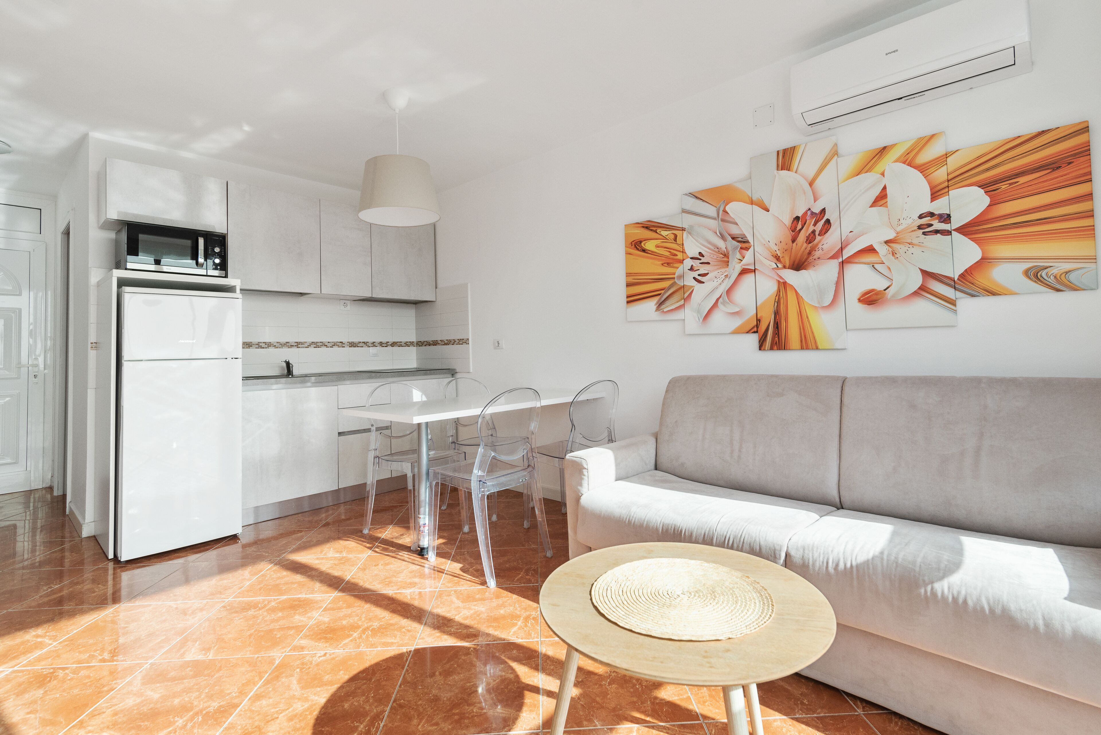 A modern kitchen with white appliances, a dining area with clear chairs, and a living area with a sofa and a wall-mounted air conditioner.
