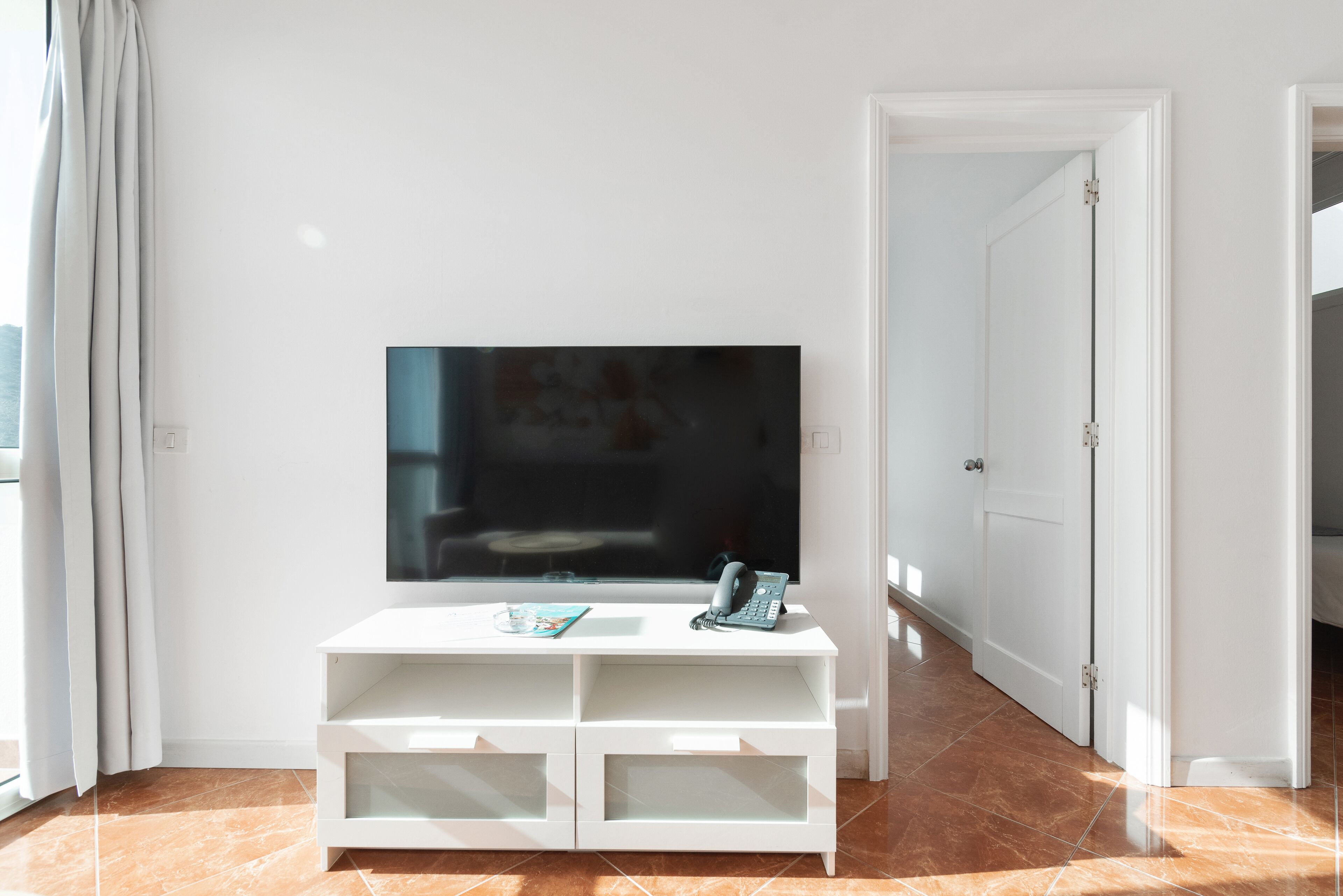 Una sala moderna con un televisor de pantalla plana sobre un mueble blanco, una puerta que conduce a otra habitación y un ventanal con cortinas.