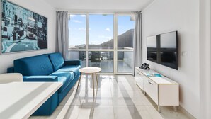A modern living room with a blue sofa, a small round table, a white TV stand, and a large window with a view of the city and hills.