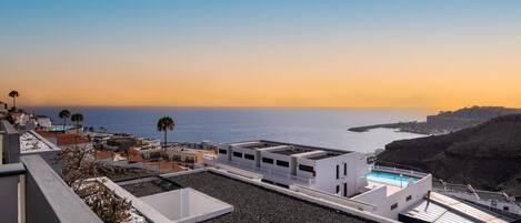 A rooftop terrace with a pool, overlooking a coastal area with buildings and a clear sky.