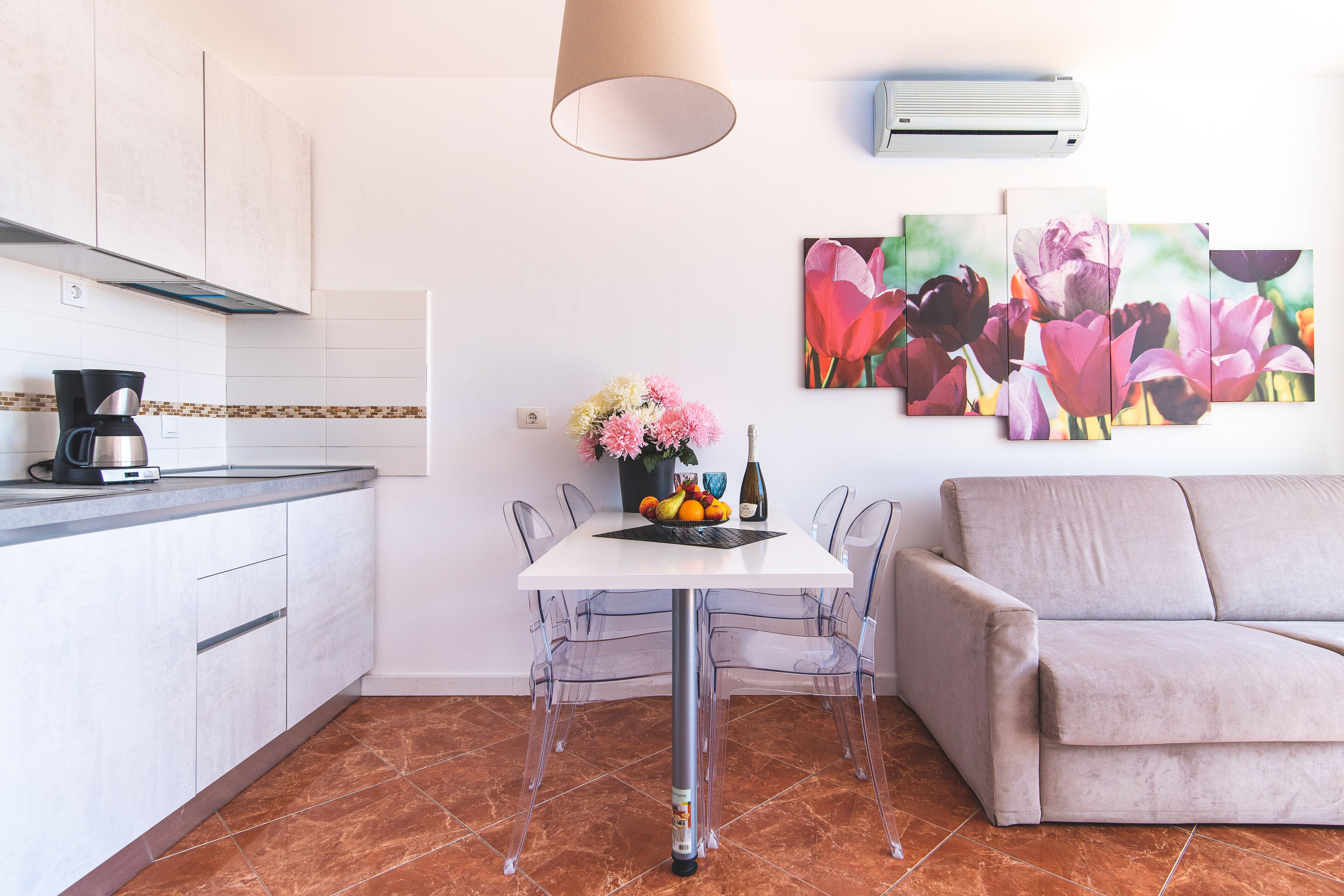 Una cocina moderna con armarios blancos, una cafetera y una mesa de comedor con sillas traslúcidas. Se aprecia un sofá y un aire acondicionado instalado en la pared.