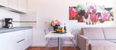 Una cocina moderna con armarios blancos, una cafetera y una mesa de comedor con sillas traslúcidas. Se aprecia un sofá y un aire acondicionado instalado en la pared.