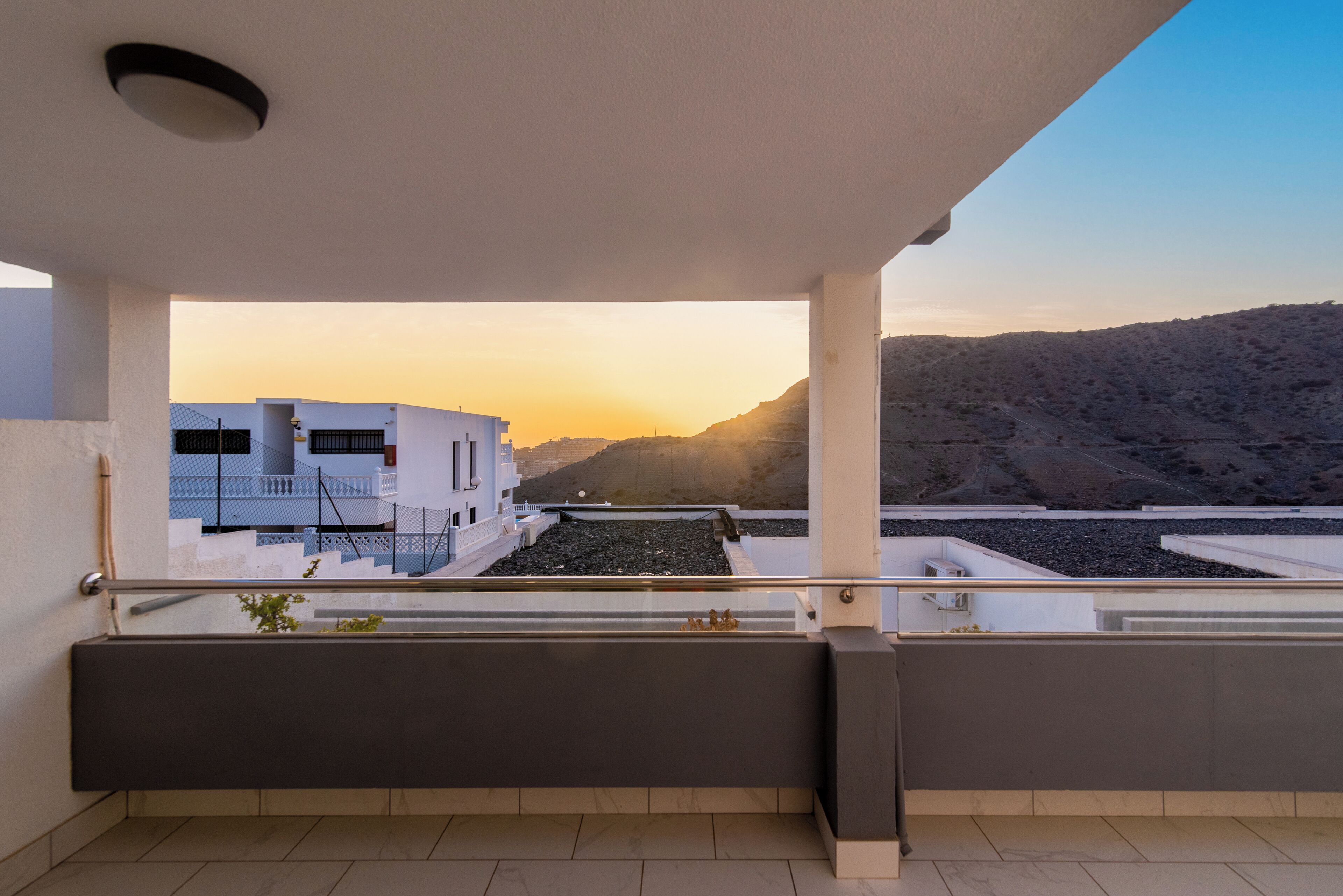 A balcony with a view of a modern building and a hilly landscape during sunset.