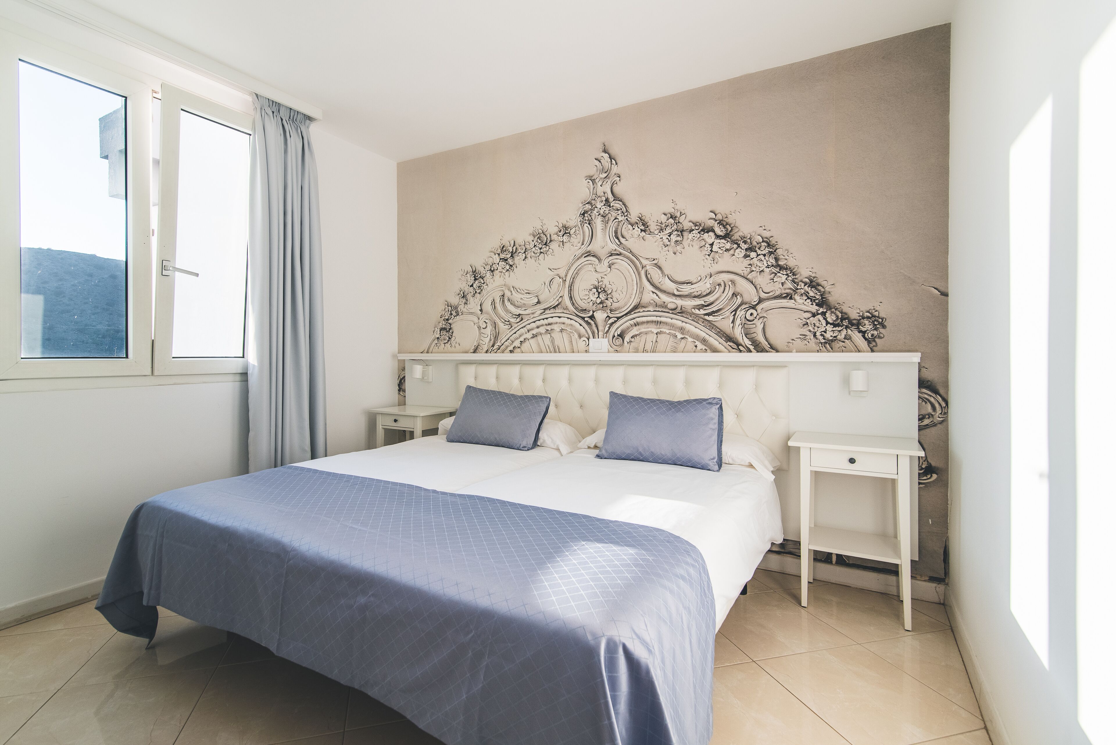 A bedroom with a large bed, two bedside tables, a headboard with a floral design, and a window with curtains.
