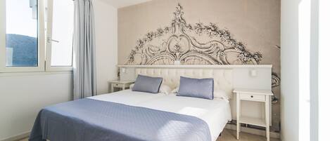 A bedroom with a large bed, two bedside tables, a headboard with a floral design, and a window with curtains.