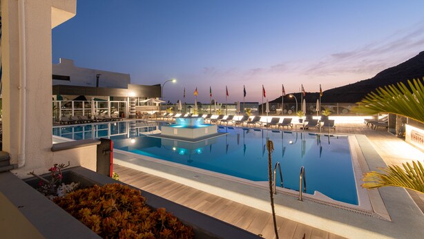 A swimming pool with lounge chairs and flags, surrounded by buildings and a mountain in the background.