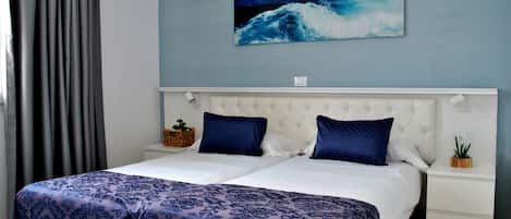 Una cama doble con cabecera blanca y dos almohadas azules, una mesita de noche con una planta y un cuadro de una ola en la pared.