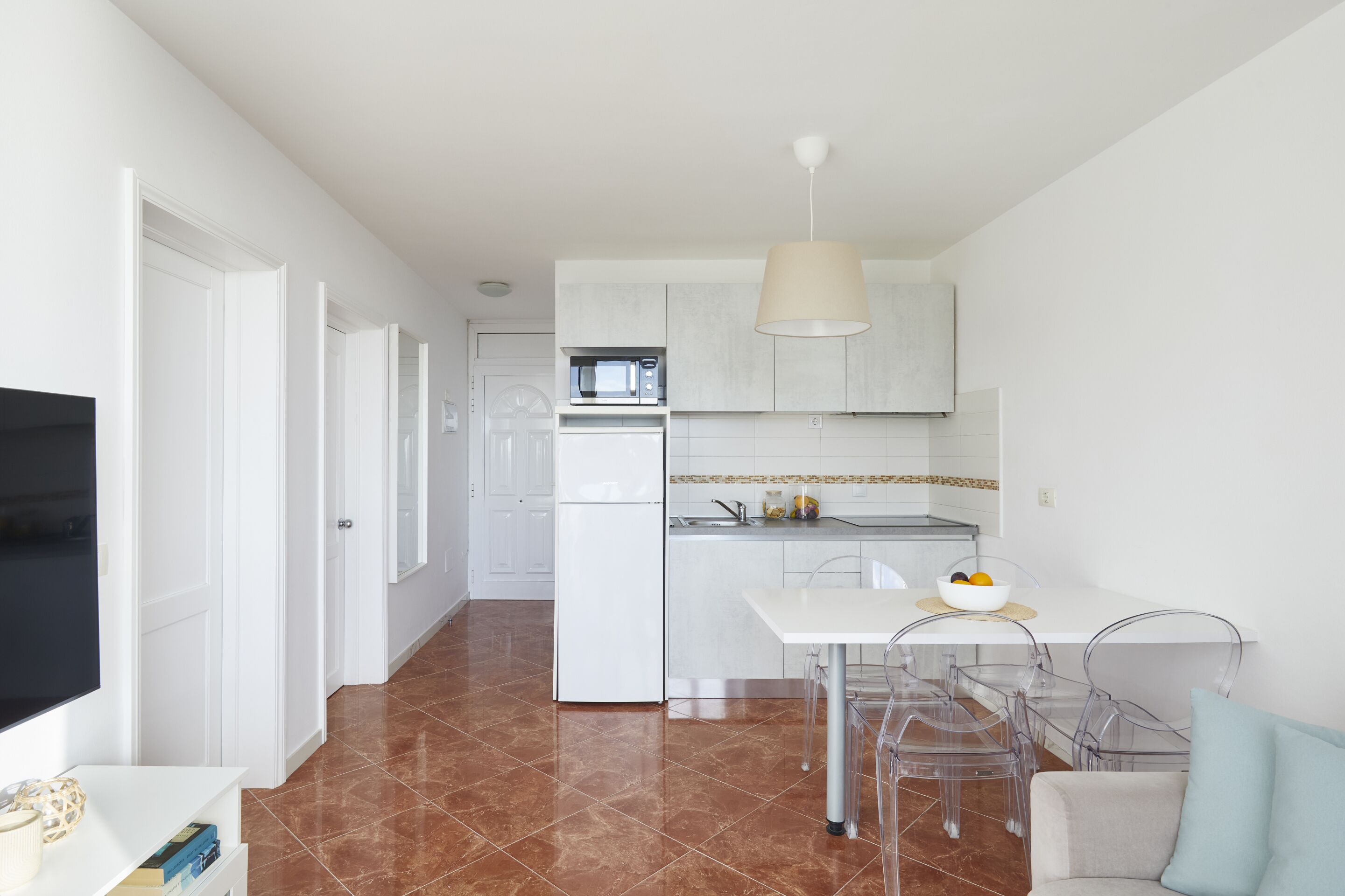 Een moderne keuken met witte kasten, een magnetron en een koelkast. Er is een eethoek met een witte tafel en transparante stoelen.