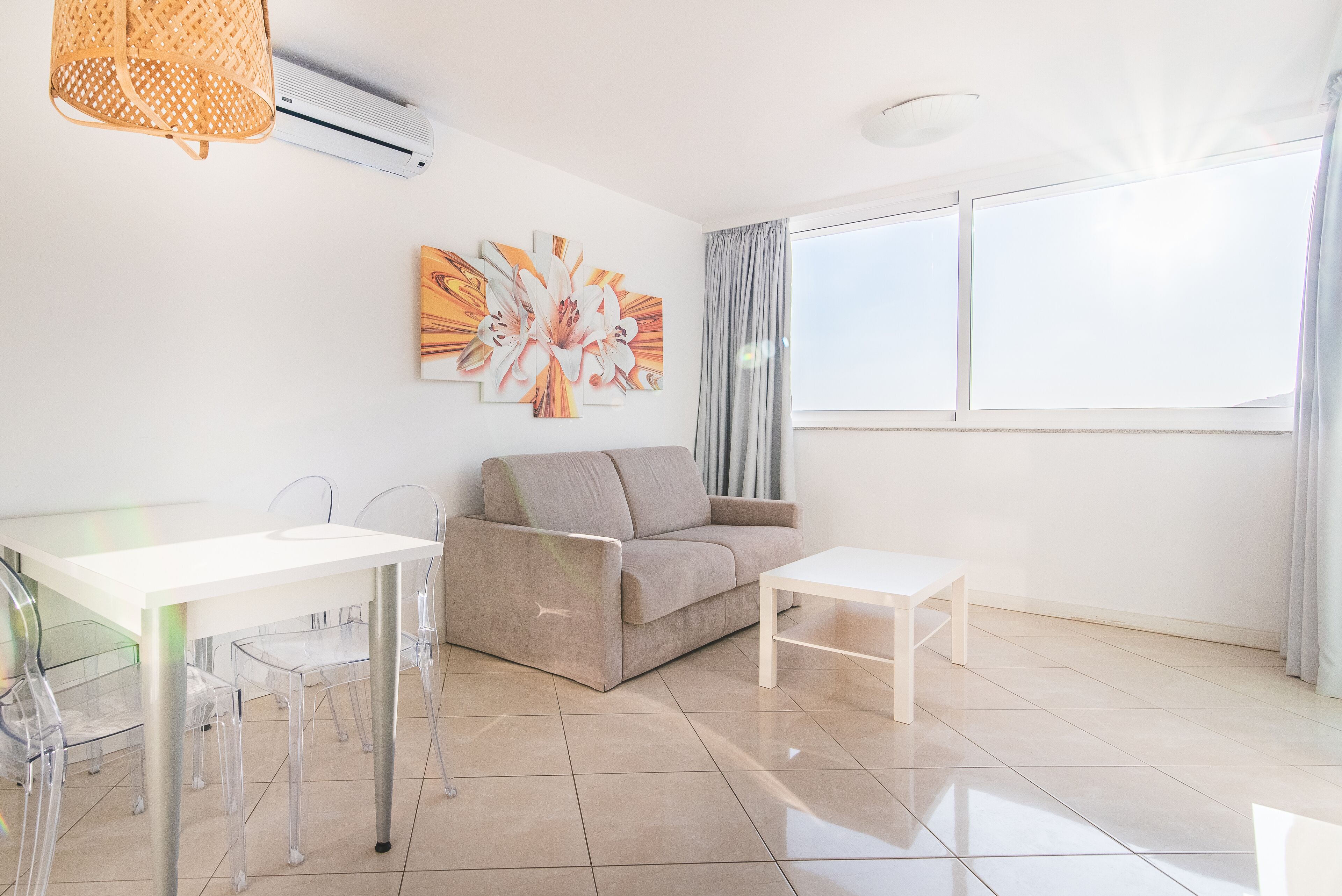 Un salon moderne avec un canapé beige, une table basse blanche et une table à manger en verre, accompagnée de chaises. On y trouve une grande fenêtre avec des rideaux, un climatiseur fixé au mur et un tableau décoratif représentant des fleurs.