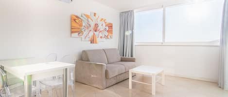 Un salon moderne avec un canapé beige, une table basse blanche et une table à manger en verre, accompagnée de chaises. On y trouve une grande fenêtre avec des rideaux, un climatiseur fixé au mur et un tableau décoratif représentant des fleurs.