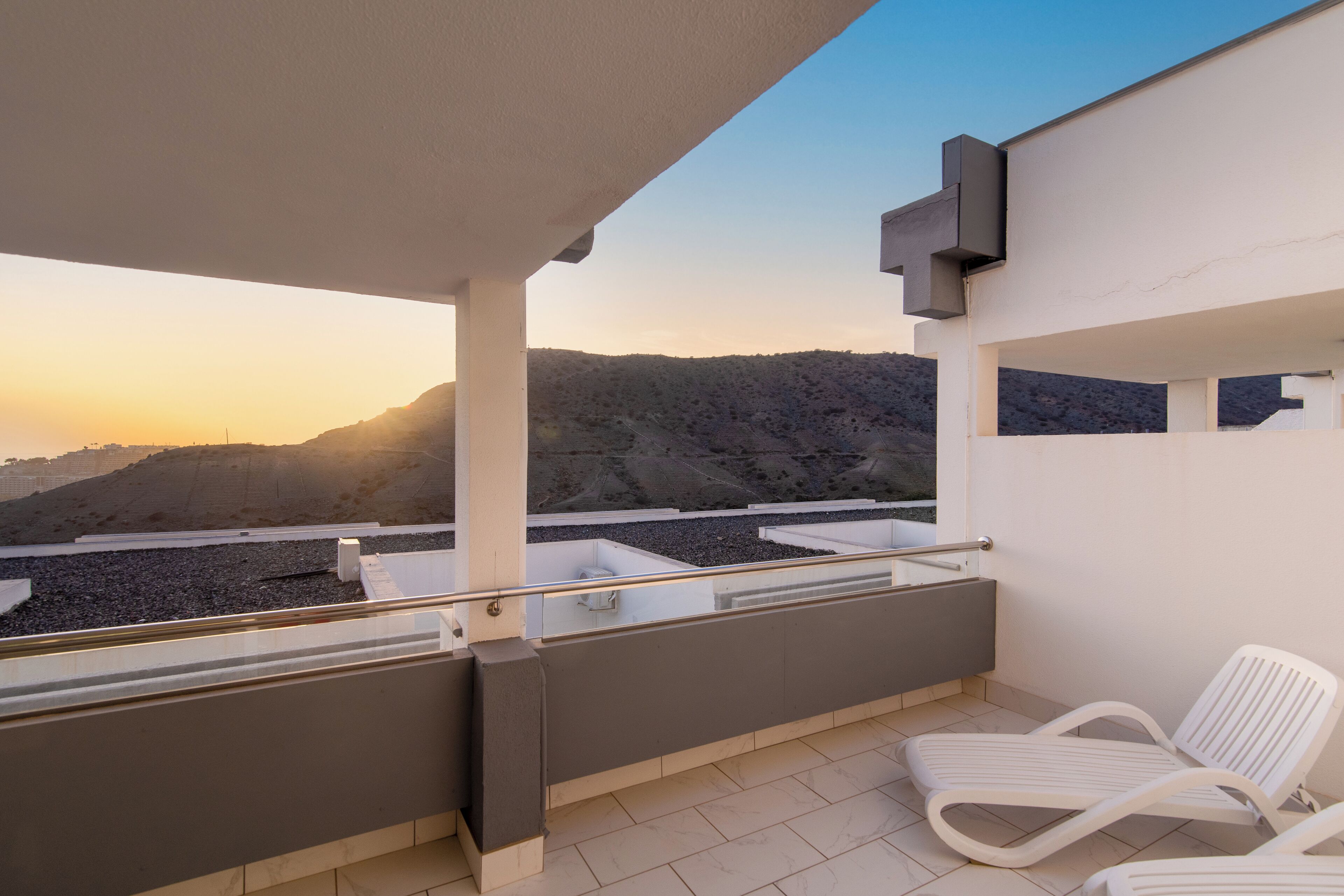 A balcony with a view of a mountainous landscape, white chairs, and a modern architectural design.